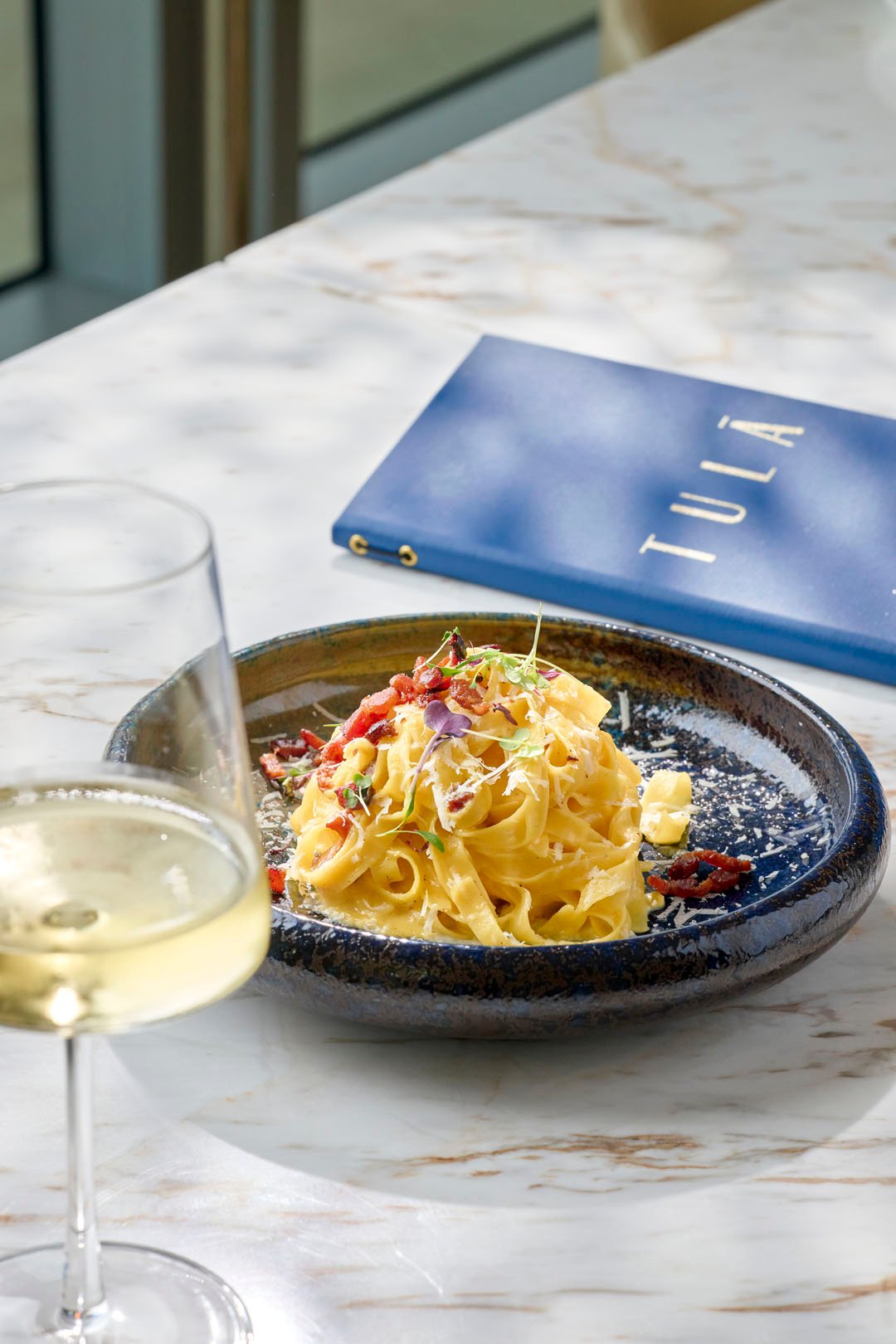 A beautiful pasta dish with micro greens in a circular black bowl with a glass of white wine at Tula
